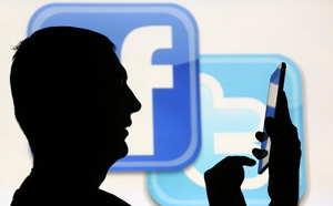 A man using a tablet is silhouetted in front of the Twitter and Facebook logos
