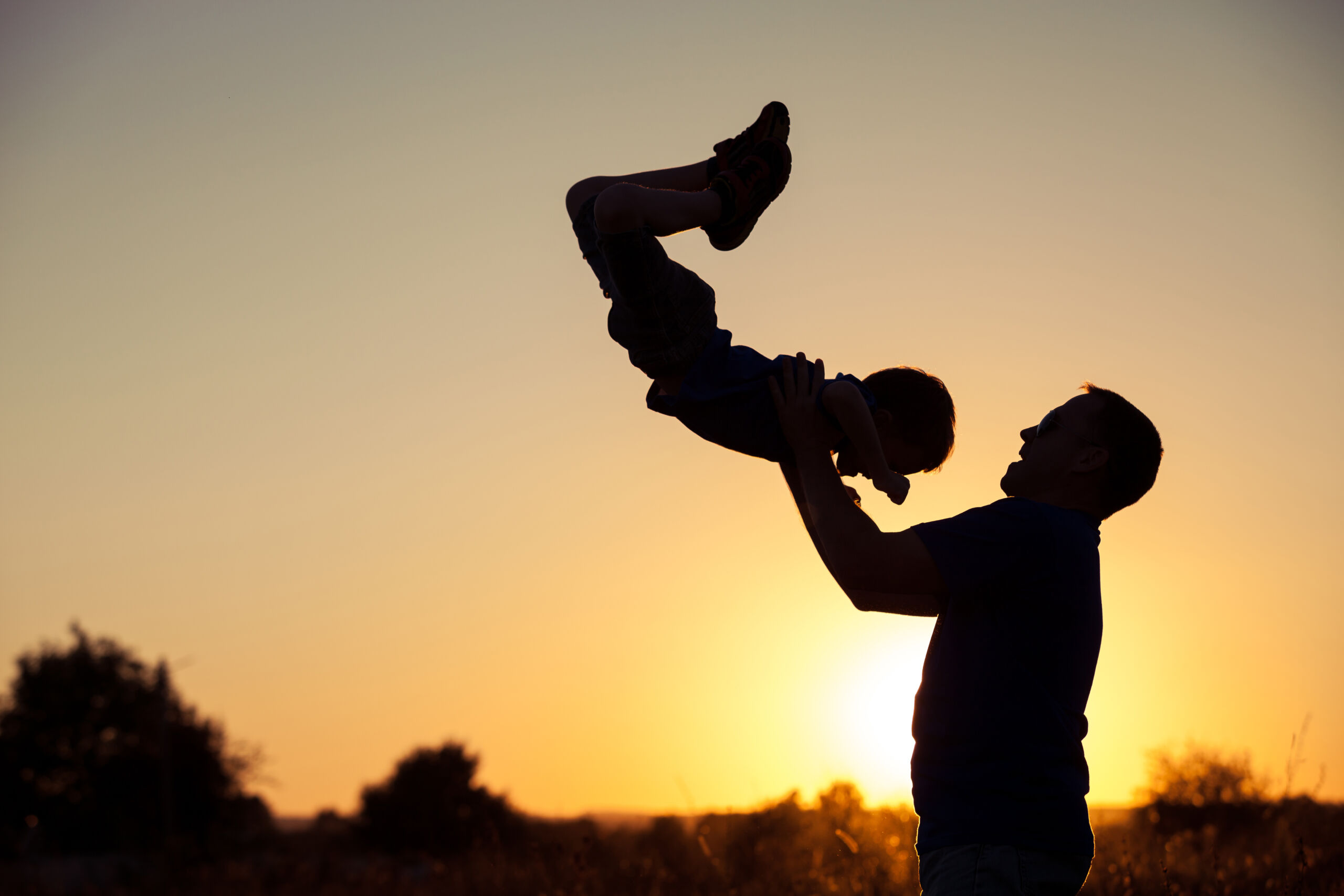 Silhouette of a dad lifting a child in the air with a sunset in the background.