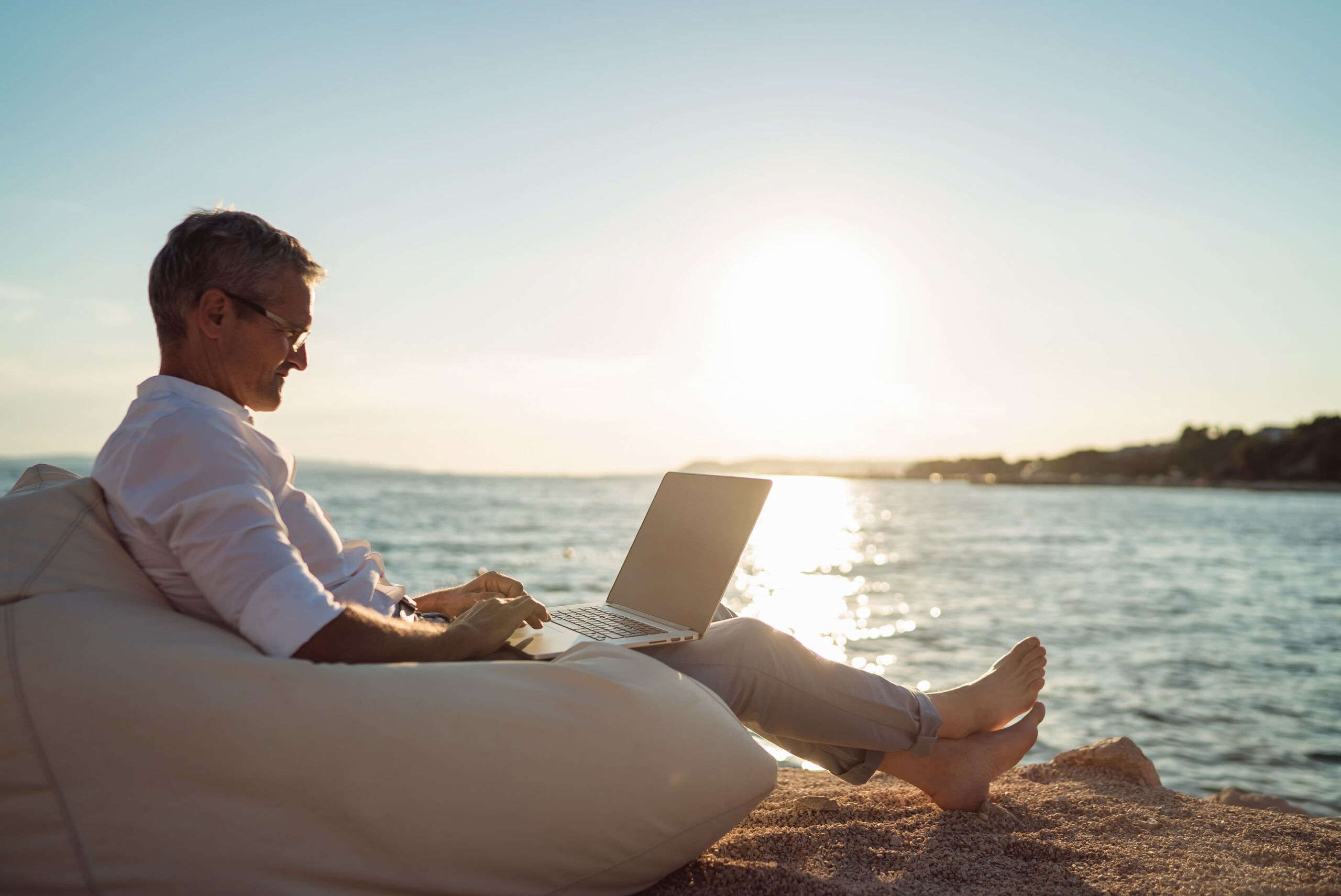 A retired man types on a laptop sitting on a bean bag on a beach next to a lake.